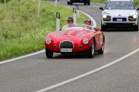 A red vintage racing car drives along green fields on a winding country road during a rally,