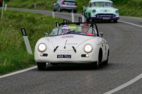 A white Porsche convertible with two occupants drives on a winding country road in front of a green