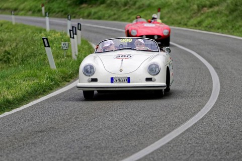 A white Porsche convertible in classic car style drives on a winding country road in front of a red
