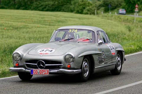 A silver Mercedes vintage car with racing stickers drives on a road in natural surroundings,