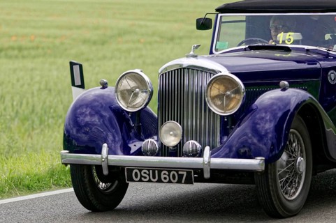 An elegant purple vintage car on a road, highlighted front headlights, surrounded by green fields,