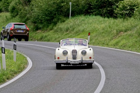 A classic convertible drives along a winding country road surrounded by green countryside, classic