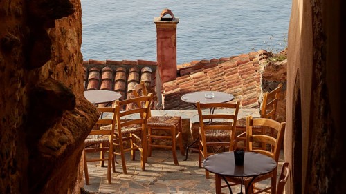 Soft morning light, narrow alley with café terrace and view of the blue sea through tiled roofs,