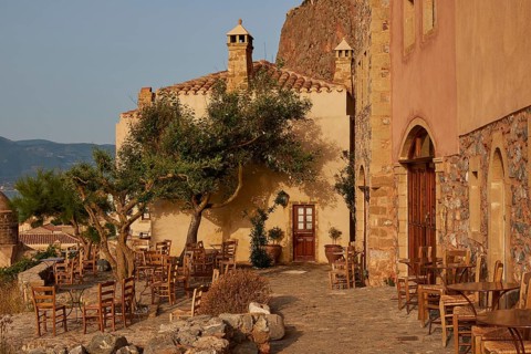 Soft morning light, a rustic outdoor café with tables and chairs on paved ground, sheltered by