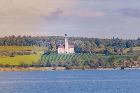 A church on a hill surrounded by vineyards and forest, with a view of a lake under a colorful