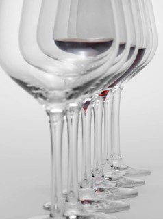 Some wine glasses standing consecutive in a row in light grey back, one partly filled with red wine