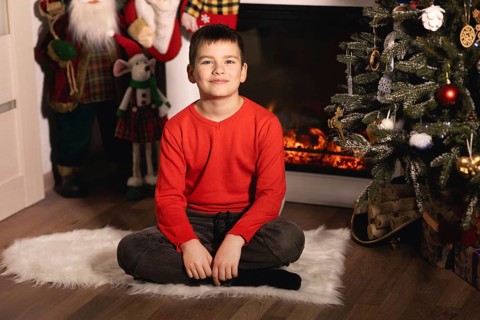 Young boy smiling, sitting on a rug by a decorated christmas tree and burning fireplace.Belarus