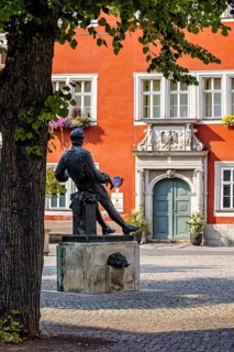 A statue in a square in front of a red, historic building with green door and blue windows, Johann