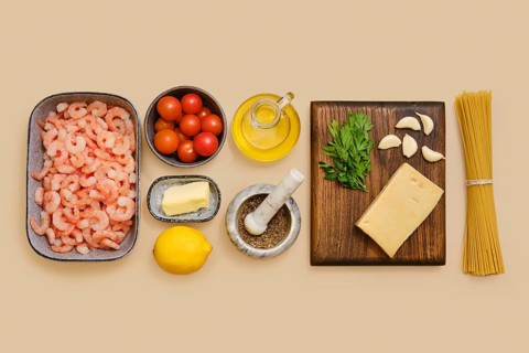 Shrimps, cherry tomatoes, garlic, and parsley lie neatly on a table along with lemon, butter, olive