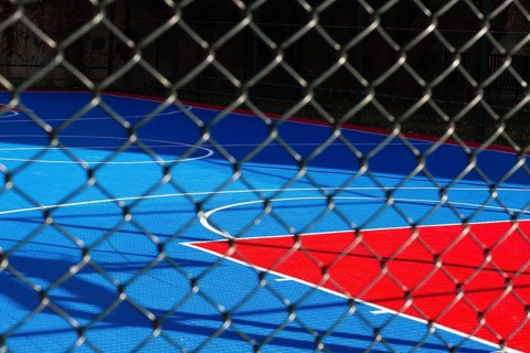 Brightly colored basketball court features blue and red sections, seen through a chain-link fence.