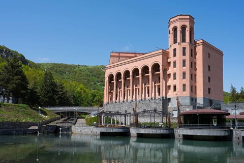 Large pink building with pillars on a river with a bridge surrounded by green hills and clear sky,