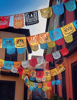 A vibrant display of colorful papel picado decorations hanging across a narrow Mexican street,