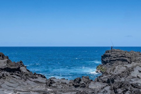 An angler stands on a massive rock on the black lava coast and looks out at the roaring Indian