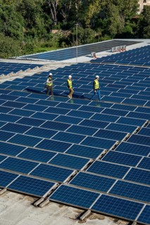 Engineers wearing safety gear are walking along rows of extensive solar panels on a rooftop,