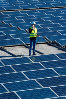 Engineer in hard hat and safety vest on commercial rooftop amid solar panels, using mobile phone
