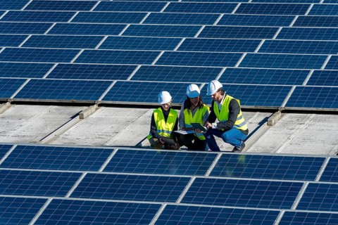 Team of engineers in hard hats and vests is discussing data on a tablet, working on a large solar