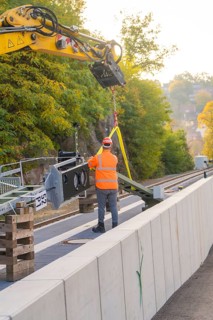 Excavator lifts objects on rails, construction worker wearing orange outfit, city in the