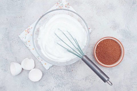 A clear bowl holds whipped egg whites, ready for dessert preparation. Next to it, a bowl of brown