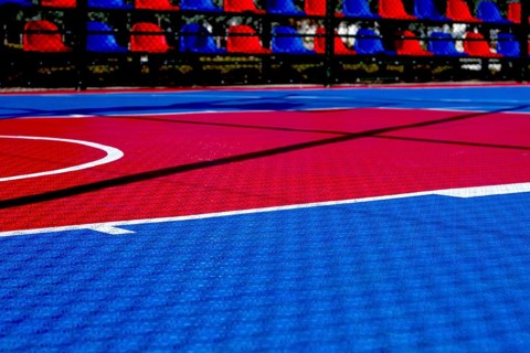 The basketball court features a striking red and blue design illuminated by bright sunlight. The