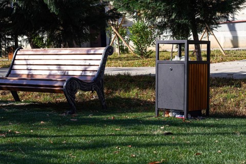 A wooden bench sits alongside a waste bin in a well-kept park. Lush green grass surrounds the