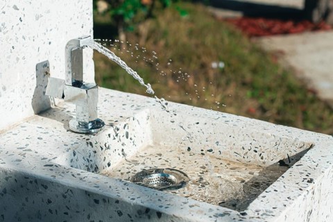 A clean tap fountain dispenses fresh water in a park surrounded by greenery. Sunlight shines on the