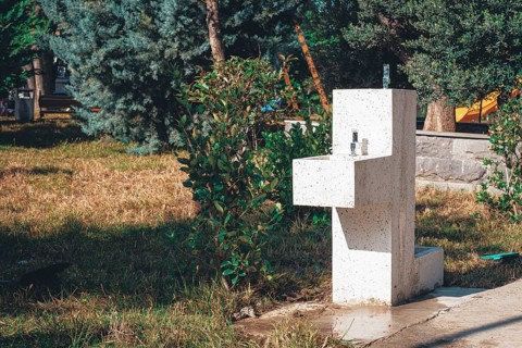 In a peaceful park, a modern water fountain is positioned among lush green plants and grass. The