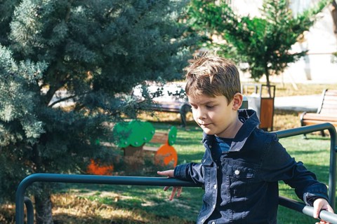 A young boy sits on a playground railing in a sunlit park, surrounded by greenery and trees. He