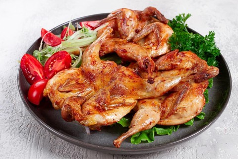Baked quail, served on a plate with crispy lettuce leaves, fresh tomatoes and herbs, on a dark