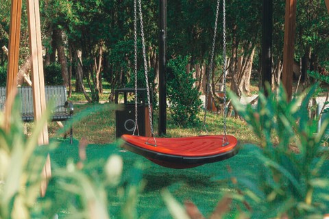 A vibrant red swing hangs suspended in a green park, surrounded by tall trees and wooden benches.
