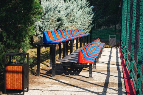Brightly colored benches line a path beside a sports field in a park. Lush greenery surrounds the