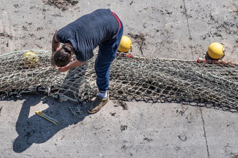 Seen from above, a fisherwoman in blue pants bends over a large fishing net spread out on the dry