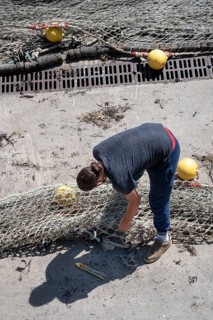 Seen from above, a fisherwoman in blue pants bends over a large fishing net spread out on the dry