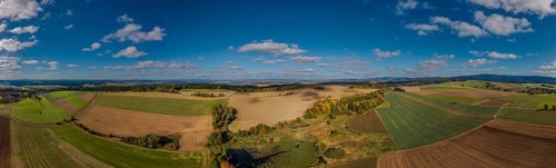 A large, open landscape shows green and brown fields with scattered trees. The sky is clear and