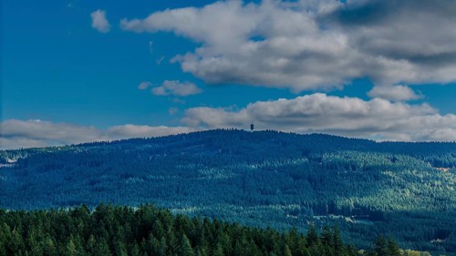 A green, wooded hill in the Bohemian Forest is visible under a blue sky with few clouds. Nature is