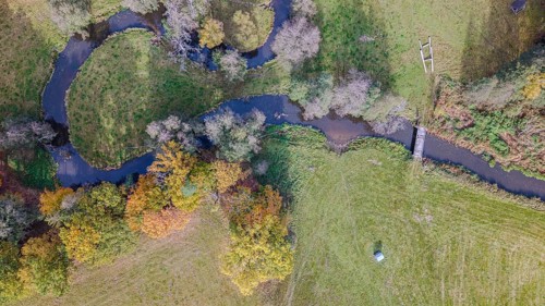 The scene shows a quiet river flowing through a picturesque meadow. Autumn colors of the trees and