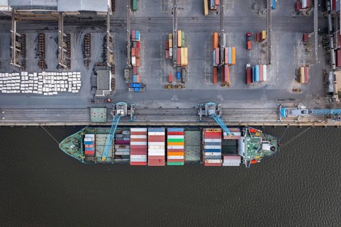 Aerial view of a cargo ship docked at a port with colorful containers and industrial surroundings.