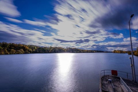 A quiet river stretches under a cloudy sky. The clouds move gently while the sunlight makes the