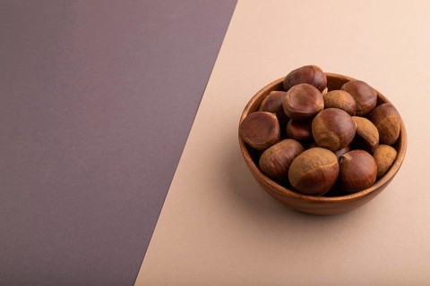 Wooden bowl with raw edible ?hestnuts on brown and gray paper pastel background, side view, copy