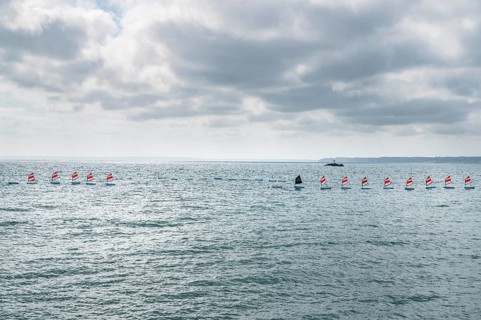 A series of small sailboats, probably optimists, with red and white ringed sails, move in formation