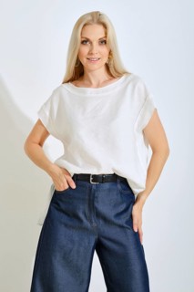 A blonde woman stands confidently in a bright studio, wearing a simple white top and loose blue