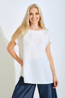 A woman stands smiling with long, light hair, dressed in a simple white top and dark denim pants.