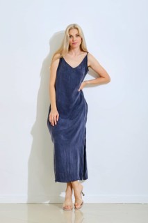 A woman stands gracefully against a plain white wall, wearing a long, sleeveless dress in a deep