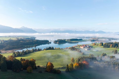 Idyllic landscape with Lake Forggensee, fog rising over green hills, farm visible in the distance,