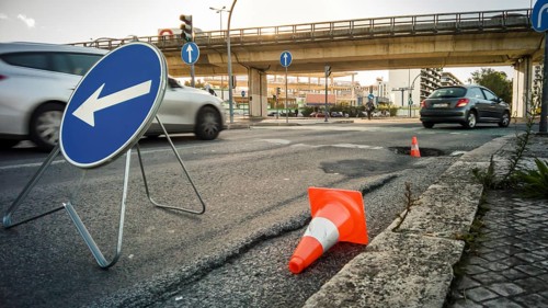 City street with large pothole marked by traffic cones and detour sign, cars passing by and