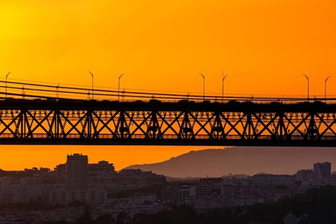 Bridge spans city skyline, silhouetted against vivid orange sunset. Soft city outlines and distant
