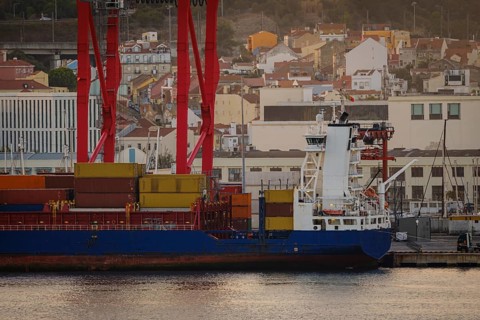 Cargo ship loaded with multicolored containers docked at Lisbon port, red cranes and city vibrant