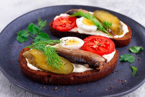 Sandwiches on rye bread with sprats, quail eggs, fresh tomatoes and pickled cucumber, on a dark