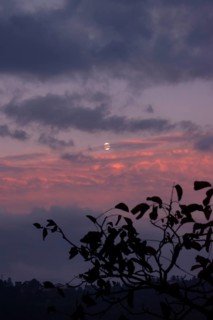 The moon shines through thick clouds in the evening sky, colored in shades of purple and pink with
