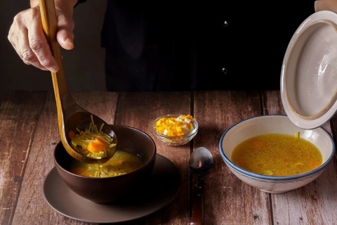 Woman serving chicken soup with a wooden spoon in a brown bowl on a wooden table