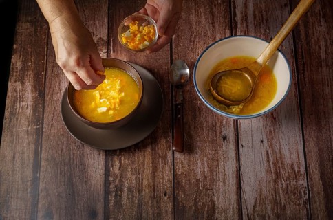 Woman pouring hard-boiled egg into a chicken noodle soup in a brown bowl on a wooden table and a